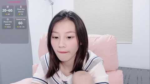 A video of an Asian woman with straight black hair, wearing a white sailor-style top, lying on a pink couch, with a digital display showing \"20-60\" in the background.