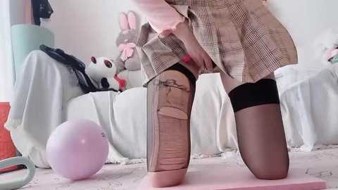 Video of a woman in a plaid skirt, black thigh-high stockings, and pink blouse, kneeling on a pink carpet, adjusting her skirt. Background features plush bunny toys and a white bed.