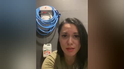 Video of a young woman with dark hair and fair skin, wearing a green top, standing in front of a blue water heater and a toilet paper roll holder.