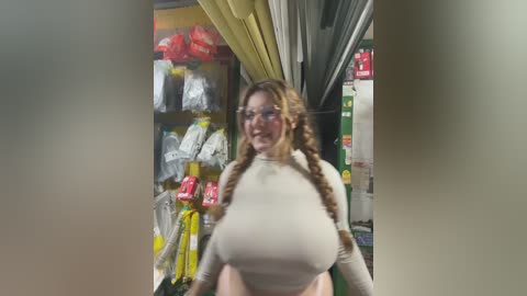 Video of a smiling woman with long braided hair, wearing a tight beige crop top, standing in a supermarket aisle filled with yellow and red packages.