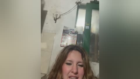 A video captures a woman with long, wavy, light brown hair smiling, standing in a dimly lit, cluttered room with beige walls, a green metal cabinet, and a bulletin board with posters.