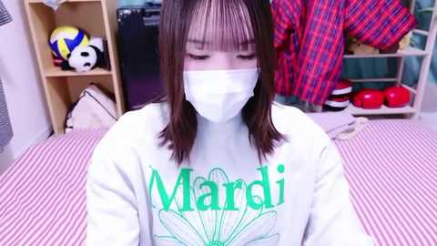 Video of a young Asian woman with straight brown hair, wearing a white face mask and a green T-shirt with \"Marci\" in green letters, sitting on a pink-striped bed in a cluttered bedroom.
