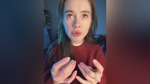 A video of a young woman with fair skin and long brown hair, wearing a red sweater, looking puzzled, with hands raised in confusion. Background shows a dimly lit room with indistinct objects.