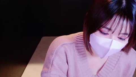 A video of an East Asian woman with straight, dark hair and bangs, wearing a white surgical mask, pink cardigan, and seated indoors against a dark background.
