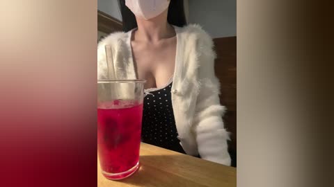 Video of an Asian woman with long black hair, wearing a white fur jacket, black polka-dot top, and a white face mask, sitting at a wooden table with a glass of bright pink liquid.