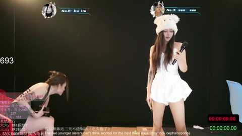 Video of a live TV show featuring an Asian woman in a white, fluffy hat and dress, holding a microphone, and another woman in a black dress and hat.