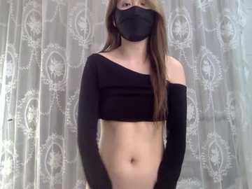 A video of a slim, light-skinned woman with long brown hair, wearing a black face mask, black off-shoulder crop top, and black gloves, standing in front of a patterned lace curtain.