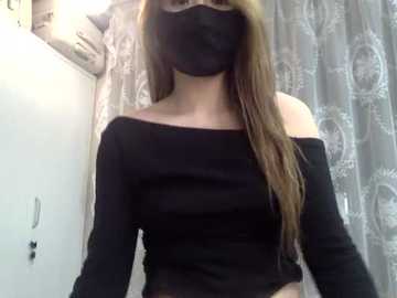 Video of a young woman with long brown hair, wearing a black face mask, black off-shoulder top, and black gloves, standing in a bathroom with a white sink and patterned shower curtain.
