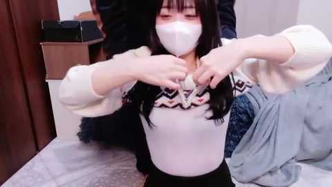 Video of an East Asian woman with straight black hair and pale skin, wearing a white face mask, a cream sweater, and black pants. She is adjusting her hair in a bedroom with a bed, blue blanket, and wooden furniture in the background.