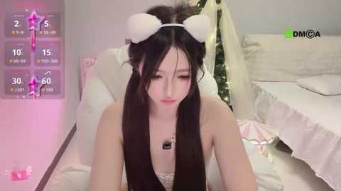 Video of a young Asian woman with long black hair wearing white rabbit ears, lying on a white bed in a pink room, displaying a \"DM\" screen.
