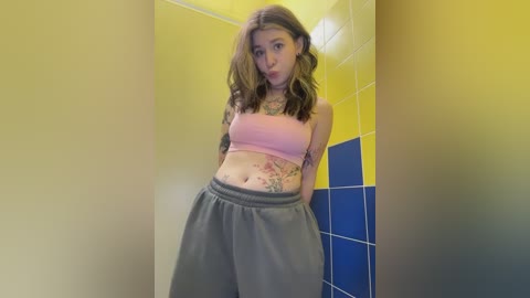 Video of a young woman with light skin, long brown hair, and tattoos, wearing a pink bandeau top and gray sweatpants, standing in a brightly lit yellow and blue tiled bathroom.