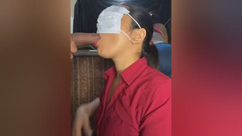 Video of a young Asian woman with light brown skin, wearing a red button-down shirt, white face mask, and white hair tie, blindfolded, leaning over a dark wooden table, mouth open, hinting at oral sex.