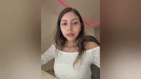 Video of a young, light-skinned Latina woman with long, dark hair, wearing an off-shoulder white lace top, sitting indoors with a pink neon ceiling light.