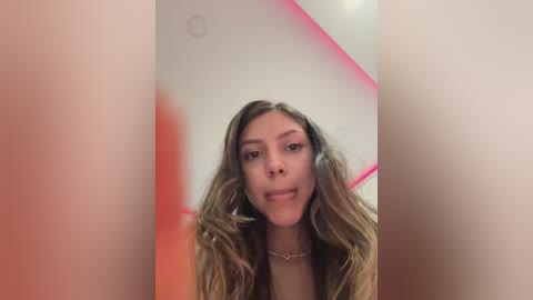 Video of a young woman with light brown skin, wavy hair, and light makeup, smiling. She's wearing a black top. Background has a red neon light and white ceiling.