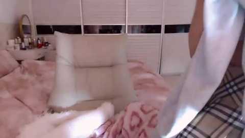 Video of a messy, pink and white bedroom with fluffy blankets, a plaid shirt, and a partially visible person in the background.