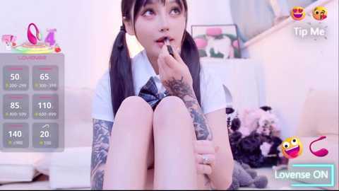 A video of a young woman with dark hair in pigtails, wearing a white shirt and denim shorts, sitting on a bed, holding her finger to her lips. The background features a plush bear and a rainbow-colored vibrator.