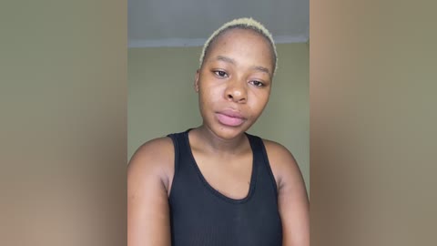 A video of a young Black woman with short, platinum blonde hair, wearing a black tank top, standing against a plain beige wall. She has a neutral expression.