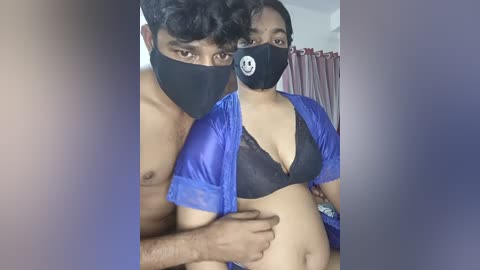 Video of a pregnant woman in a blue top and black bra, wearing a face mask, standing next to a shirtless man with short hair. Background includes a bed with purple curtains.