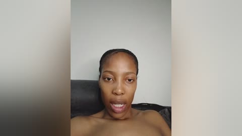 Video of a topless, dark-skinned woman with short hair, mouth open in a surprised expression, reclining on a black leather couch against a plain, light-colored wall.