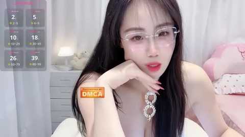 A video of an Asian woman with long black hair, wearing glasses and a necklace, lying on a bed in a pink room, with a digital calendar overlay.