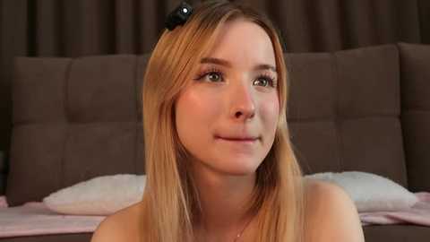 Video of a young, fair-skinned woman with straight blonde hair, wearing a black headband, smiling slightly, sitting on a bed with a brown headboard and white pillows.