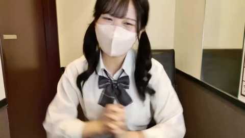 A video of an Asian woman with long black pigtails, wearing a white face mask and a school uniform, sitting at a desk in a beige-walled room.