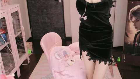 Media: Video of a woman in a black, lace-trimmed mini dress kneeling beside a pink, plush baby chair. The room features a white shelf with toys, a dark grey wall, and a large poster on the right.