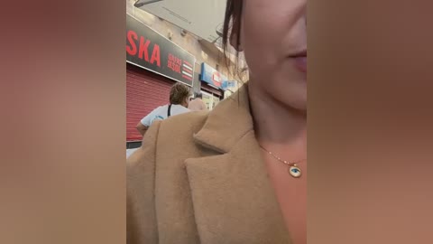 Video of a woman in a brown coat, holding a bag, walking towards a closed red \"SKA\" store in a dimly lit mall.