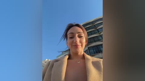 Video of a smiling, fair-skinned woman with brown hair, wearing a beige coat, standing in front of a modern building with balconies, under a clear blue sky.
