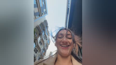 Video of a smiling woman with long brown hair, wearing a beige jacket, standing in front of a tall, narrow alley between two multi-story buildings with balconies.