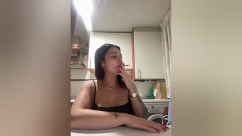 Video of a woman with short black hair, medium build, in a small kitchen, wearing a black tank top, sitting on a counter, holding her chin, with a small tattoo on her wrist.