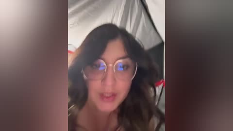 Media: Video of a woman with wavy black hair, wearing glasses, and a red top, captured in a blurry, low-light setting, with a white tent-like structure in the background.