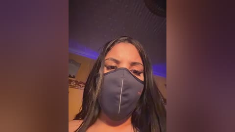 Video of a woman with long, straight black hair and medium brown skin wearing a dark blue surgical mask indoors. Background features a ceiling with recessed lighting and a blurred, warm-toned room.