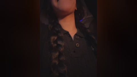 A video of a person in a black, button-up shirt adorned with a fur trim on the collar, standing in a dimly lit room with dark, blurry background. The focus is on the person's upper body, neck, and part of the face.