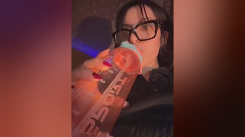 Video of a woman with short black hair, glasses, and a black jacket drinking from a large orange Gatorade bottle. Background is blurry with a reddish tint, suggesting a dimly lit setting.