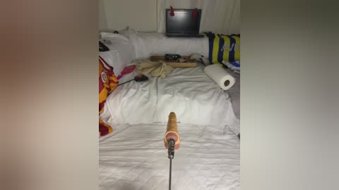 A video of a messy, cluttered bedroom with white sheets, a rolled-up sleeping bag, a flat-screen TV, and a colorful blanket. A wooden paint roller handle protrudes from the foreground.