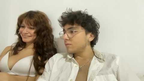 Video of a smiling, middle-aged woman with long brown hair in a white bra, and a young man with curly black hair and glasses, wearing a white shirt, seated against a plain white background.