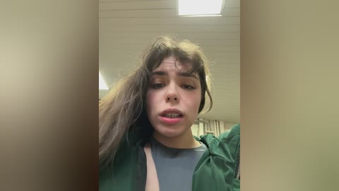 A video of a young woman with long brown hair, wearing a green jacket and gray shirt, looking distressed, standing in a dimly lit hallway with beige walls and fluorescent lighting.