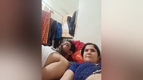 A video shows a man and woman lying on a bed in a cluttered room with hanging clothes and cardboard boxes in the background.