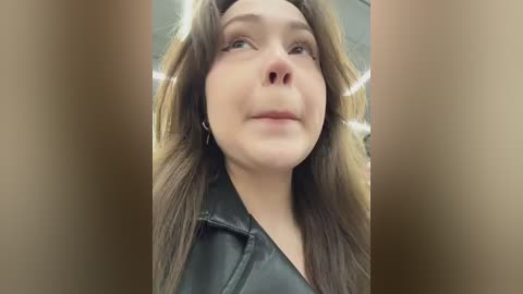 Video of a young woman with long brown hair, fair skin, and light makeup, wearing a black leather jacket, looking upward with a neutral expression. Background is blurred, possibly a room with soft lighting.