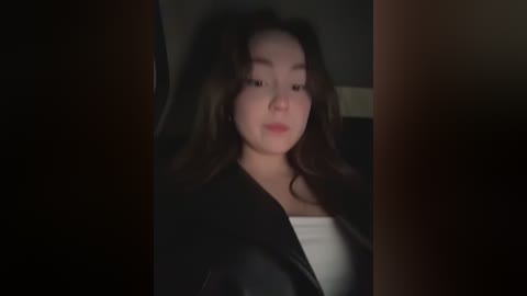 A dimly lit video of a young Asian woman with long, wavy dark hair, wearing a black leather jacket over a white top, standing in a dark, possibly indoor setting.