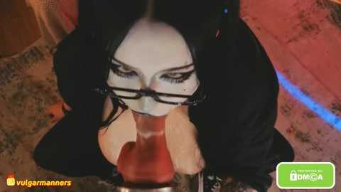 A video of a young woman with pale skin, long black hair, and glasses, kneeling, performing oral sex on a man's erect penis. The background features a red and blue carpet.