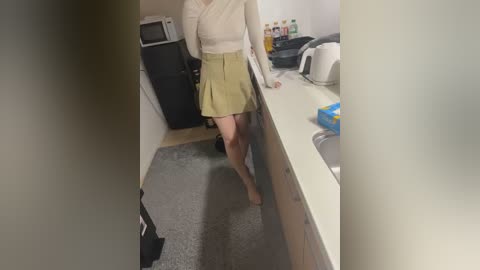 Video of a woman with light skin and a slim build, wearing a beige pleated skirt and white long-sleeved blouse, leaning against a kitchen counter in a dimly lit room.