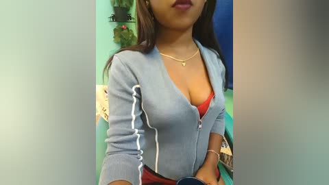 Video of a young woman with medium brown skin, wearing a light blue zip-up jacket over a red bra, standing indoors. Background includes green walls, shelves with plants, and a blue garment.