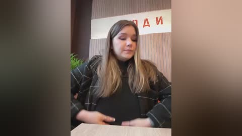 Video of a young woman with long, straight, light brown hair, wearing a black turtleneck and plaid jacket, seated at a desk. Background features a potted plant and partially visible \"DAM\" sign.