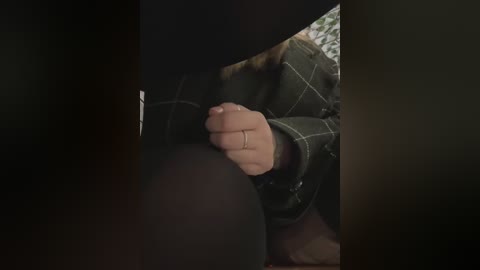 A video of a woman's foot, adorned with a ring, peeping through a dark opening. She wears a black plaid shirt, with her arm resting on a white-patterned surface.