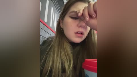 A video shows a young woman with long, straight, light brown hair, wearing a black jacket, wiping her eye with a red hand, in a dimly lit, brick-wall cafe setting.