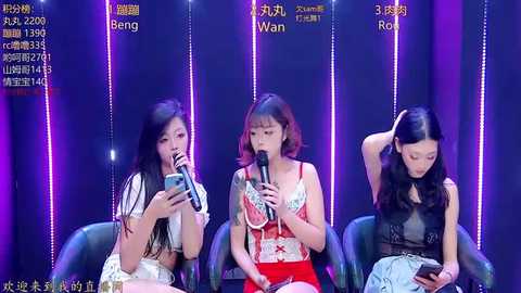 Video of three young Asian women with long hair, seated in black chairs against a purple-lit backdrop, singing into microphones, wearing casual and elegant outfits, on a stage.