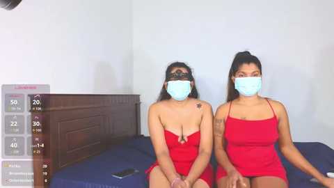 Video of two women in red tank tops, masks, and face shields, sitting on a blue bed, with a dark wooden headboard and white wall background.