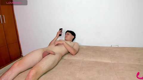 A nude man with fair skin and short black hair lies on a beige carpet, taking a selfie with his smartphone. The background features a white wall and wooden cabinet.